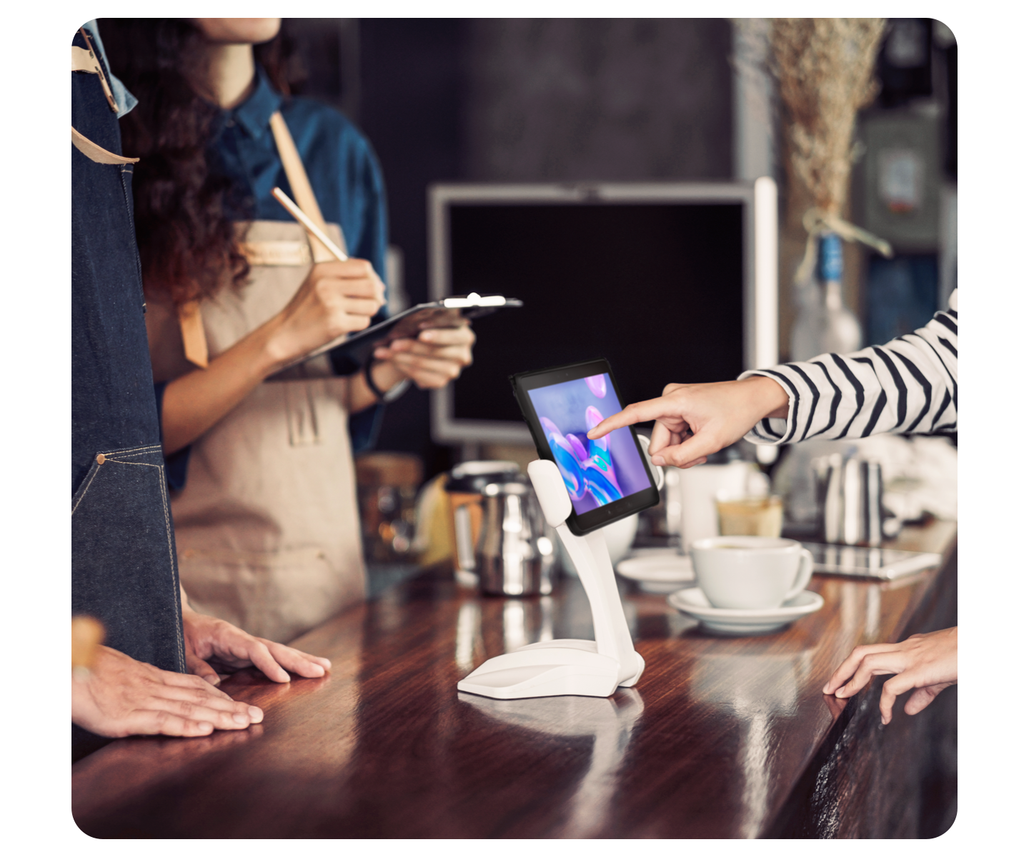 A white Samsung tablet on a counter is used to pay for a coffee. The torso of a man and of a woman are pictured on the left.