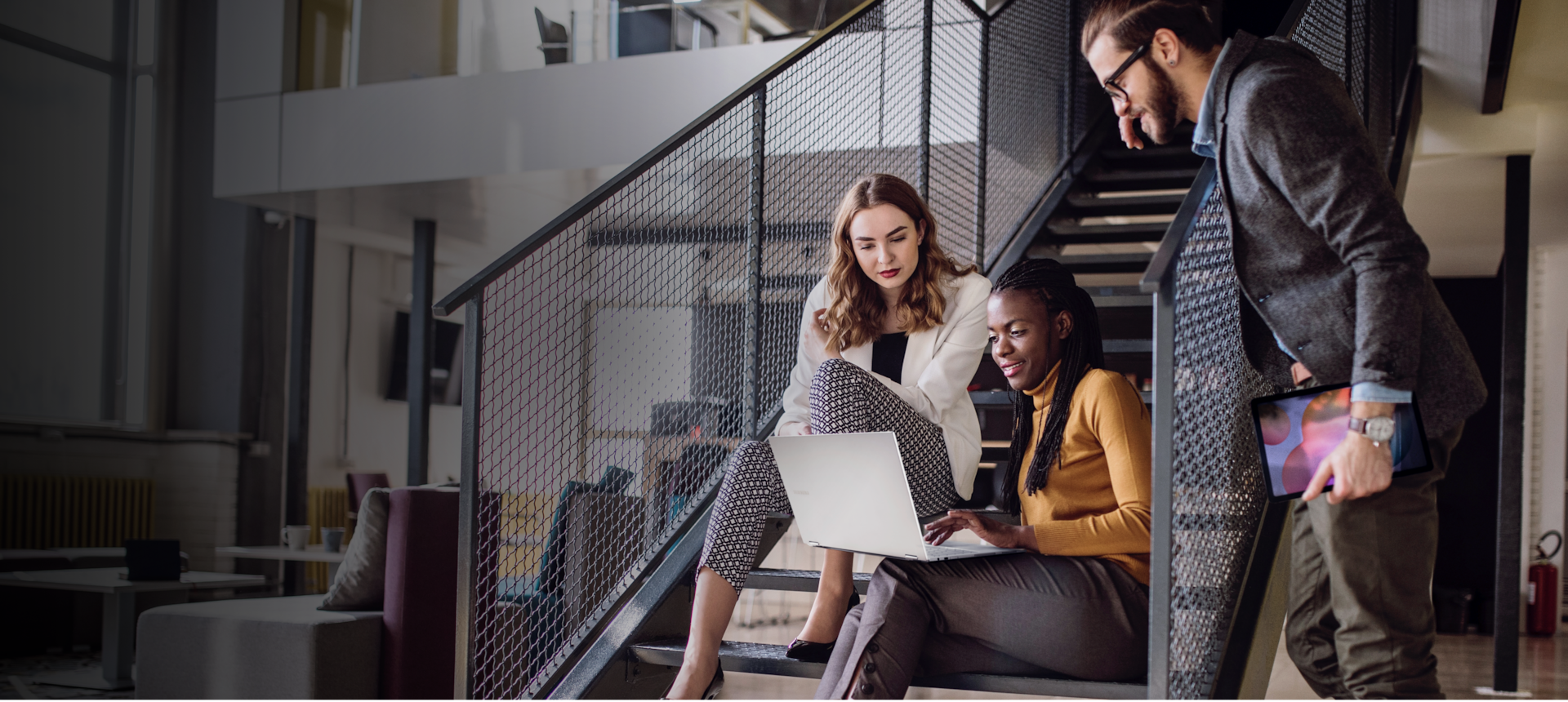 Two women sit on a staircase looking at a Samsung laptop together. A man leans on the staircase holding a Samsung tablet.
