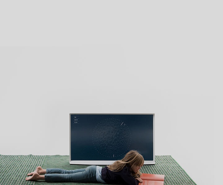 A young girl is laying on a green area rug reading a book. Next to her, The Frame has been placed directly on the rug without a stand.