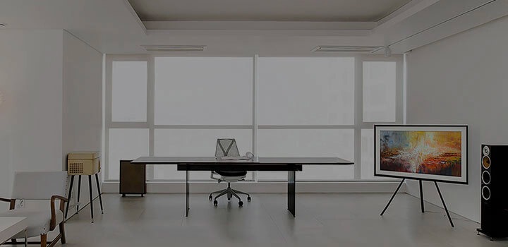 The Frame TV and several appliances are inside a bright, white modern room with large windows. The Frame TV has been placed on the right side of the room on a studio stand. It is showing a piece of artwork on its display.