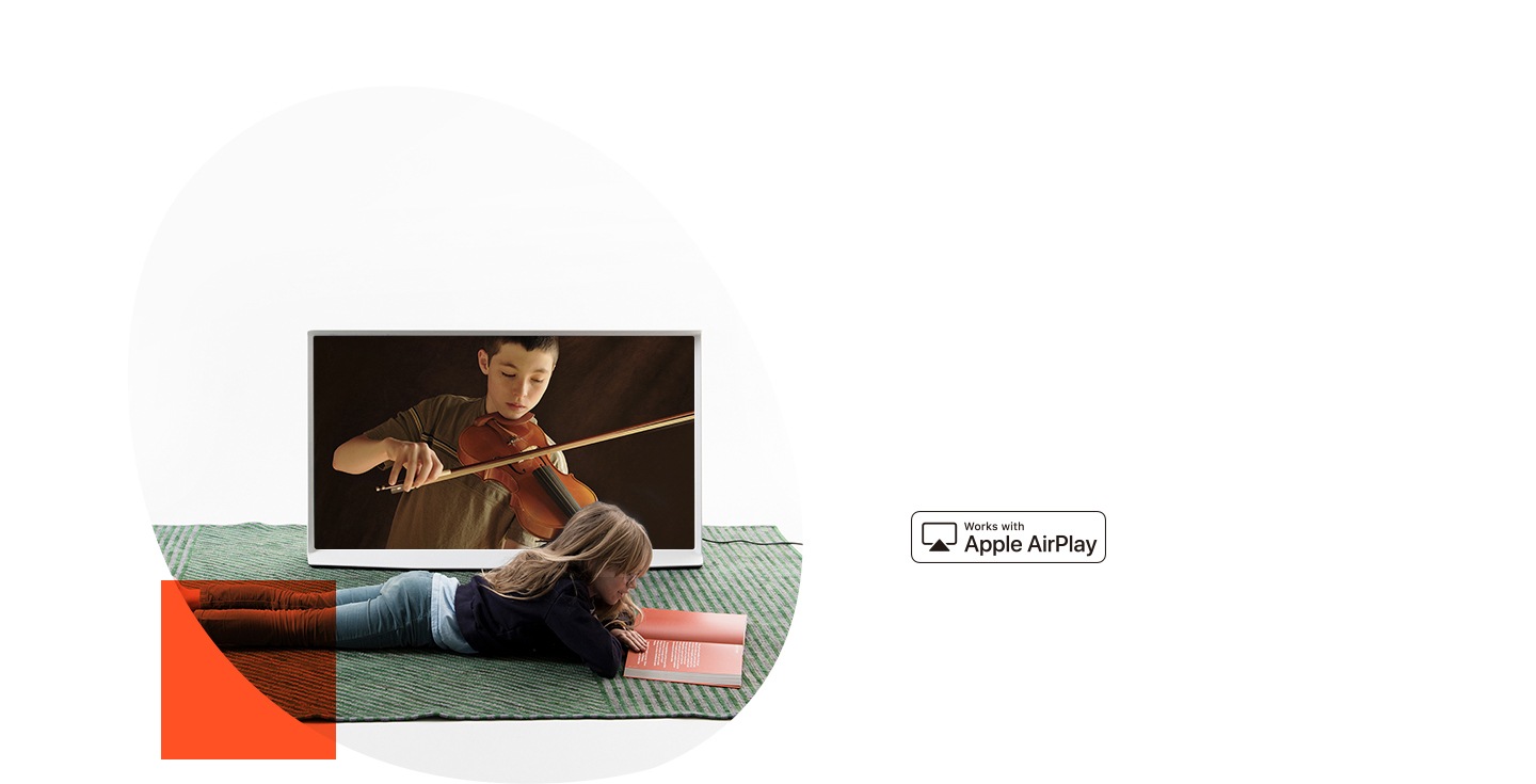 Little girl reads a book on the floor in front of a White The Serif that is connected to an AirPlay to show a boy playing a violin on-screen.