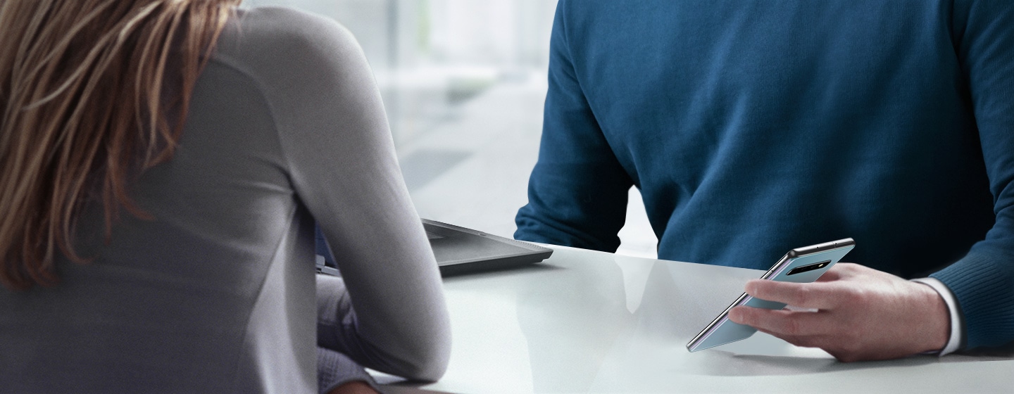 Image of a man and a woman sitting across a table from one another. The man is holding Galaxy S10 plus in his hand.