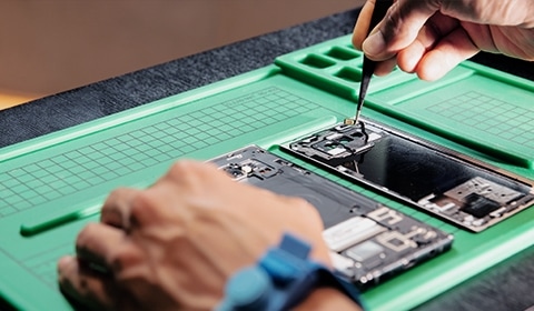 Closeup of the hands of a technician repairing a phone on a table. The phone has been taken apart with the interior components exposed