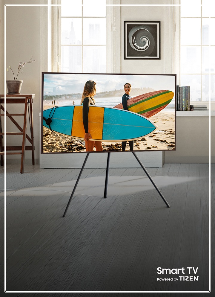 Model Frame televizora u crnoj boji postavljen je na stalak u kutu prostorije, a na zaslonu je otvoreno Samsung Smart Hub sučelje, a odabir je postavljen na izborniku usluge Netflix. Frame televizor izravno otvara ugrađenu aplikaciju usluge Netflix i pokreće prikazivanje filma koji pruža usluga Netflix: 6 Underground. U kutu zaslona prikazuje se tekst da je ovaj pametni televizor pokretan operativnim sustavom TIZEN.