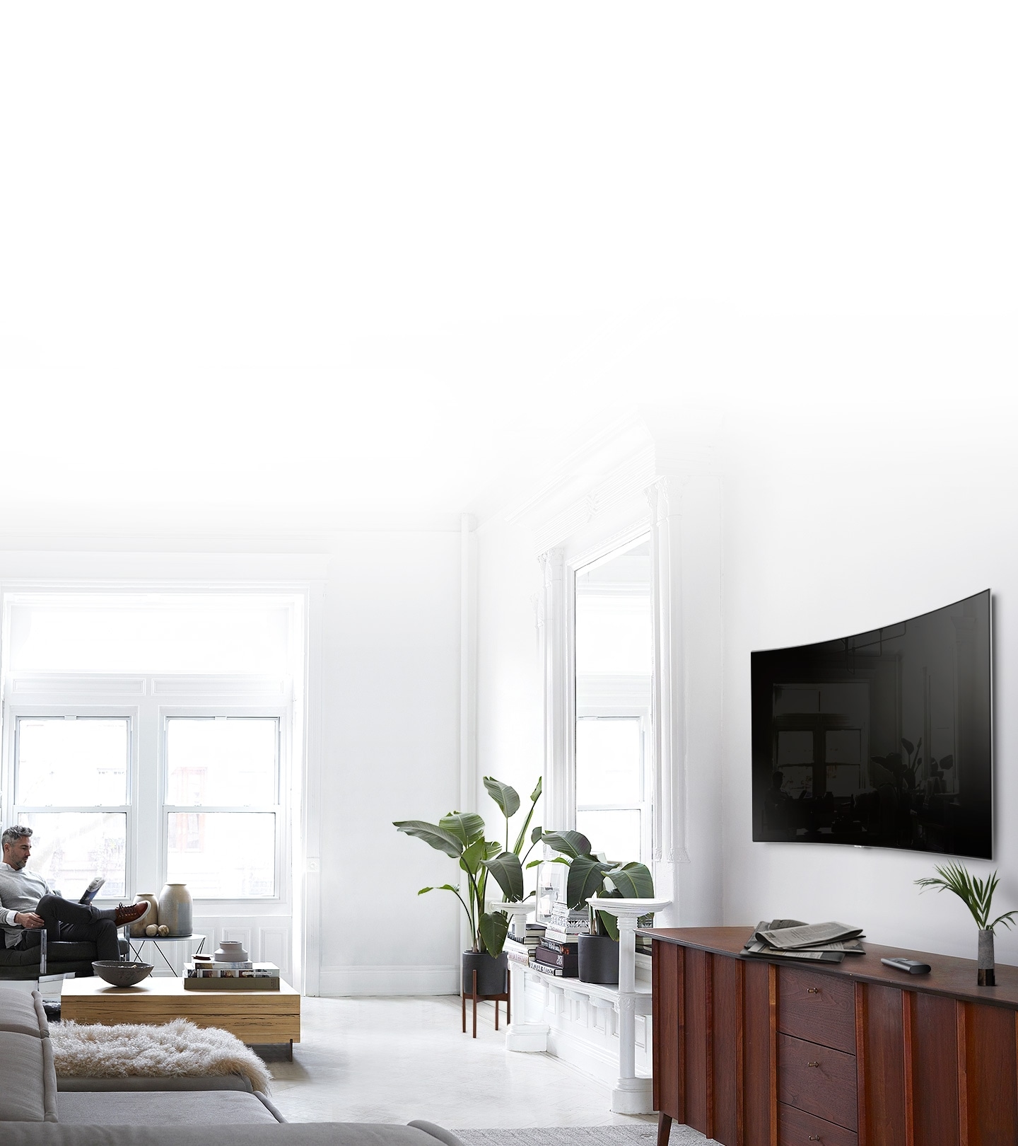 A kissé oldal nézetből látható nappaliban a fehér árnyalatok dominálnak, a QLED TV szilárdan a falra rögzítve látható. Egy férfi az ablak előtt ül egy székben és újságot olvas.