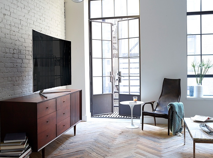 A QLED TV egy barna faasztalra helyezve. Vele szemben egy köralakú asztal és egy szék látható. A kép a nappali és az ívelt TV képernyő harmóniáját mutatja be.