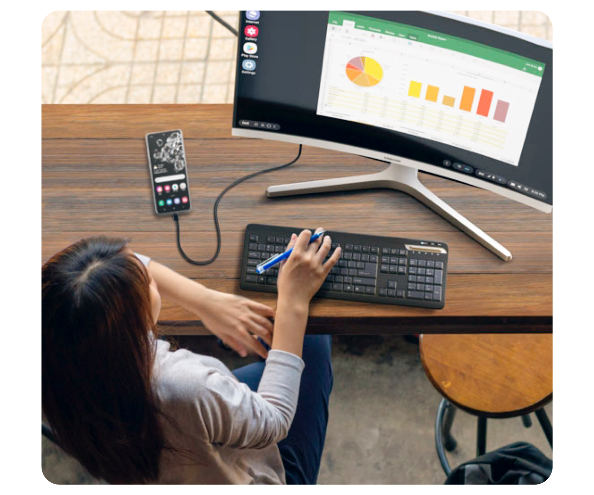 Shown from above, a woman sits at a desk with a Samsung curved monitor, a black wireless keyboard and a black smartphone.