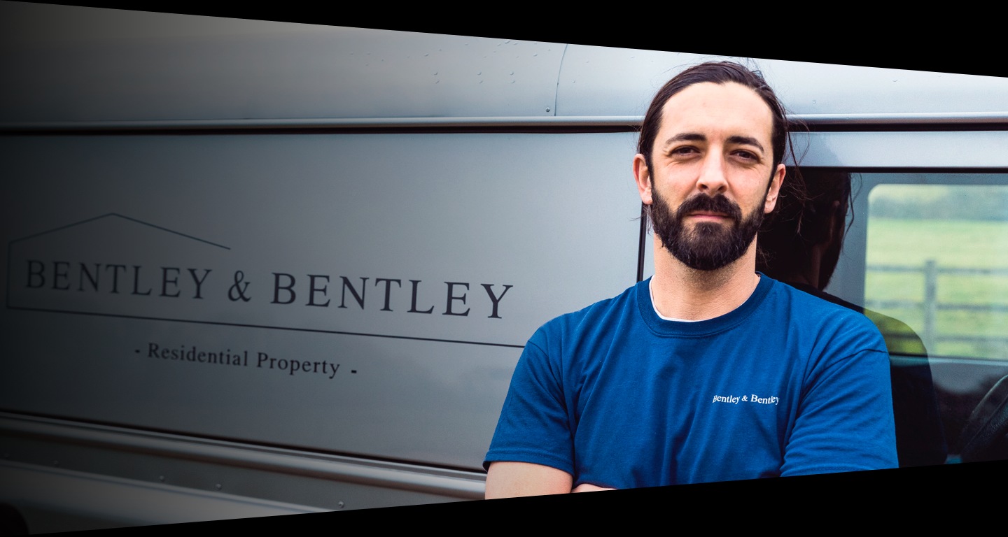A man with a beard wearing a blue t-shirt leans on a silver van behind him while looking at the camera and crossing his arms.