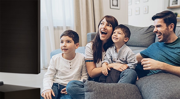 A laughing and smiling family of four are sitting closely together on a sofa enjoying a big screen TV.