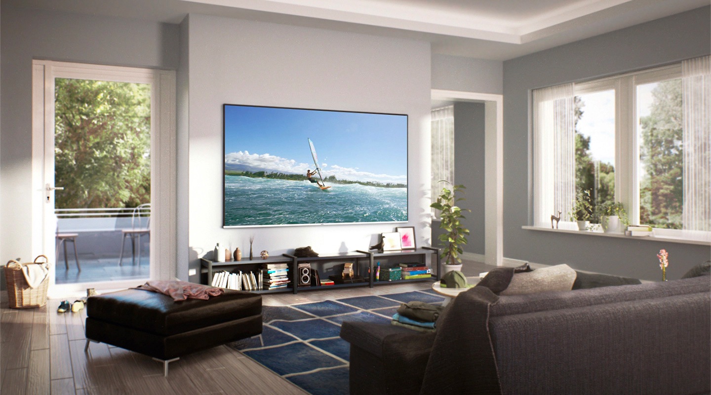 Wide view of cozy living room with daylight from out of the window. A Super Big TV is hung on the wall. On the screen, a man is enjoying surfing.
