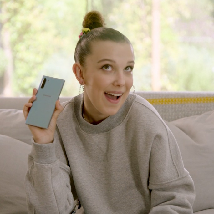 Millie Bobby Brown sitting on a couch, holding up an Aura Glow Galaxy Note10