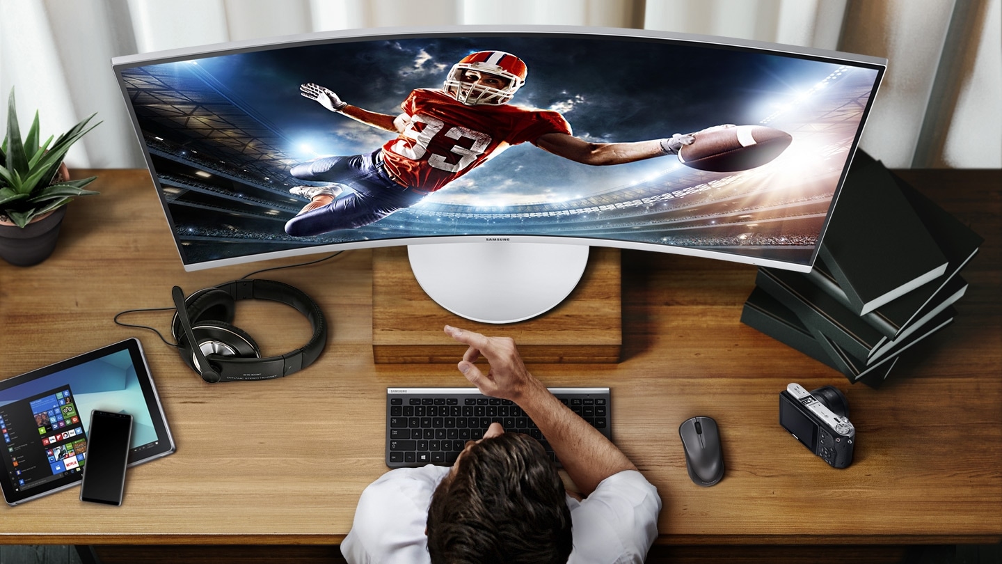 There is a monitor on the room table, and rugby sport is shown on the monitor screen. The man is staring at the monitor, and on the table are tablets, cell phones, headphones, mice, keyboards, cameras, potted plants, and books.