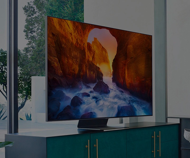 Un TV The Frame è appeso alla parete avorio di una stanza; accanto un'ampia finestra affaccia su un paesaggio con alberi e edifici in lontananza. Il The Frame Samsung è posizionato su un supporto e montato su un mobile turchese. Sullo schermo viene mostrato un paesaggio di alcune formazioni rocciose.
