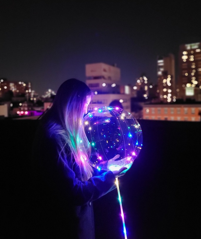 Summer night photography of a woman holding a glowing LED balloon while bokeh lights glow in the background