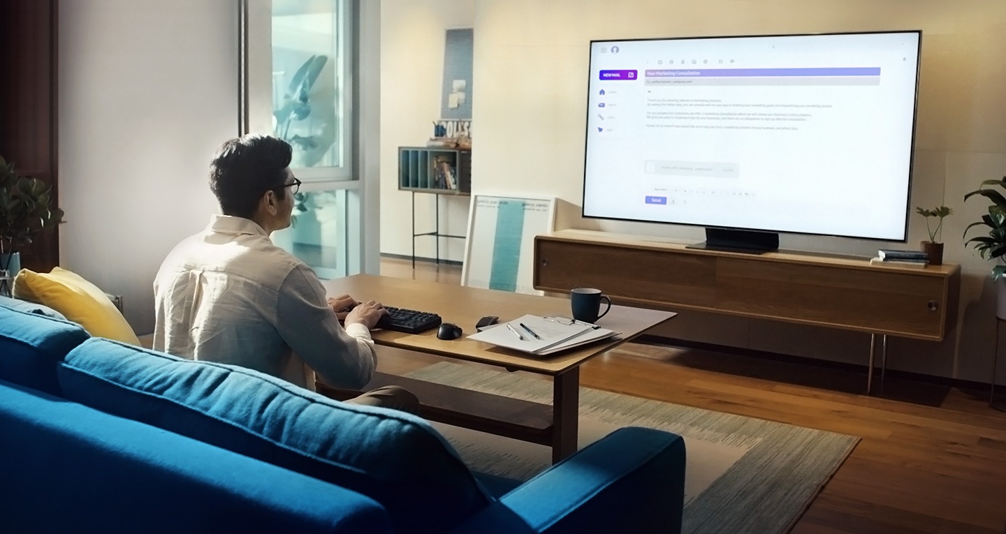 A man is sitting on a couch in his living room typing on a keyboard that's connected to the Samsung Smart TV which displays what's on his computer back at the office via Remote Access. He's writing an email which is shown on the screen.