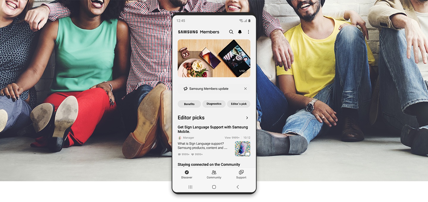 Personas de diversas razas sentadas en el piso y riendo. Un teléfono inteligente Galaxy en primer plano está ejecutando la aplicación Samsung Members.