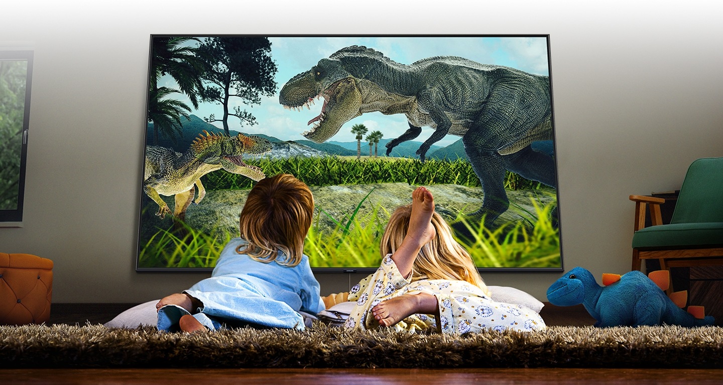 2 kids are watching a movie with lying down on the lug. The movie lively shows two dinosaurs fight.