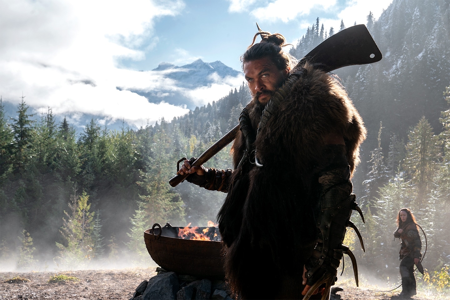 A man in the mountains stands with a weapon in front of a fire pit with a woman behind him.