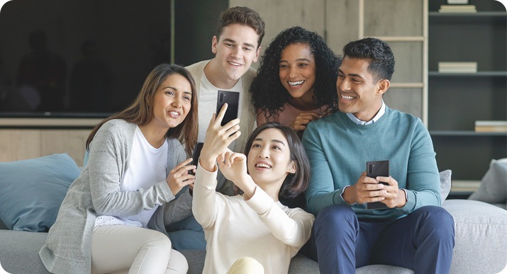 A group of friends are sitting on the couch while the woman in the middle is crouching on the floor and holding her smartphone up to take a selfie of the group.