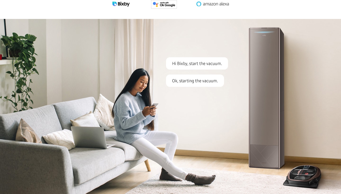A woman sits on her sofa with her smartphone. There is a smart air conditioner and a smart robot vacuum in the room. 2 conversation bubbles appear near the woman with the text "Hi Bixby, start the vacuum" and "Ok, starting the vacuum". 3 AI assistant logos are displayed above: Bixby, Hey Google, and Amazon Alexa.