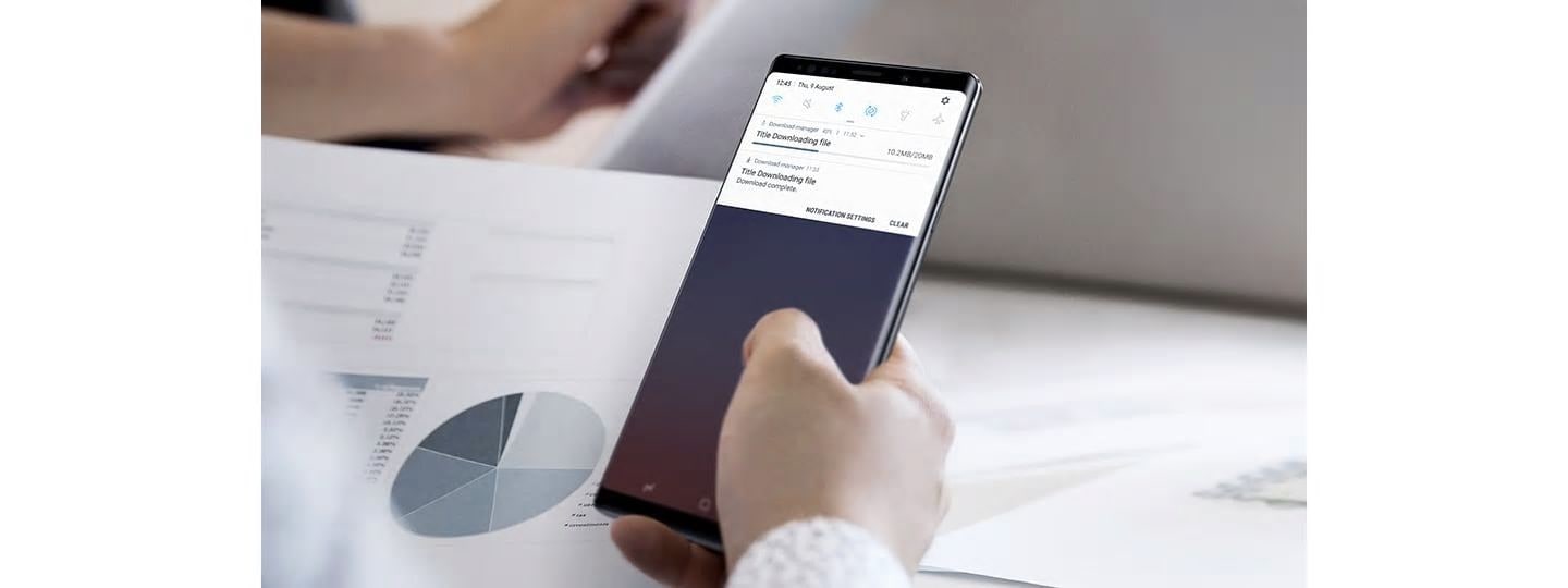 Image of a person holding Galaxy Note9 with the download GUI onscreen, one download is finishing and another is completed