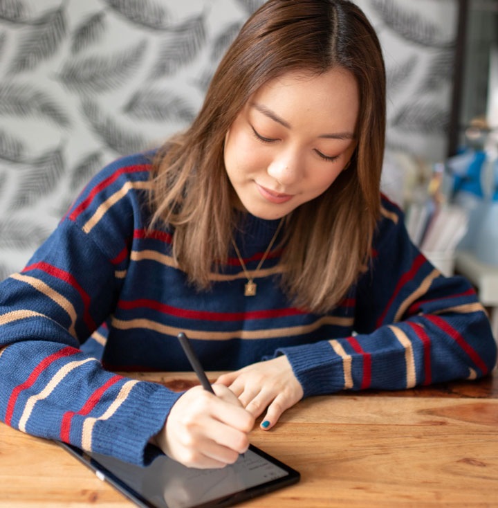 Amanda Rach Lee bullet journaling on her Galaxy Tab S6 Lite with a dark grey S Pen as she sits at a wooden desk. She's wearing a navy blue sweater with red and pink stripes.