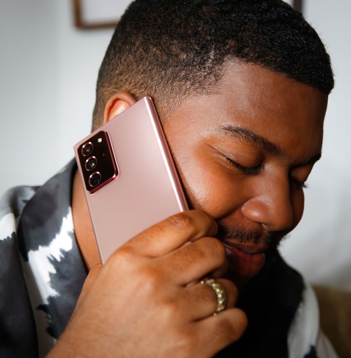 Khalid holds a Mystic Bronze Galaxy Note20 up to his right ear and closes his eyes, smiling. He's sitting on a beige couch and wearing a black and white tie dye t-shirt.