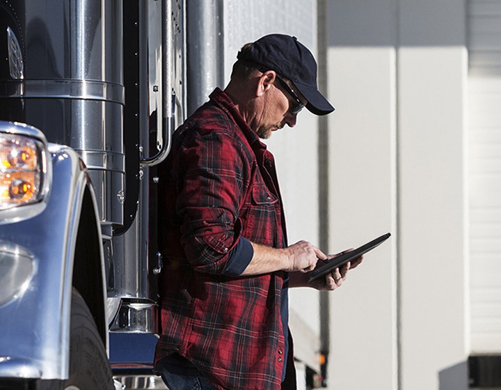 A truck driver leaning on his truck while looking into a Samsung tablet