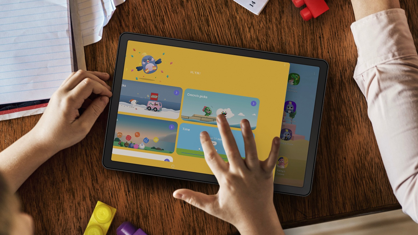 Una tablet sobre una mesa rodeada de papeles y bloques de LEGO. Las manos de un niño están cerca y su mano derecha toca la pantalla de la tablet. En la pantalla está la interfaz del usuario de la aplicación LEGO, que muestra diferentes tipos de entretenimiento que tu hijo puede disfrutar.