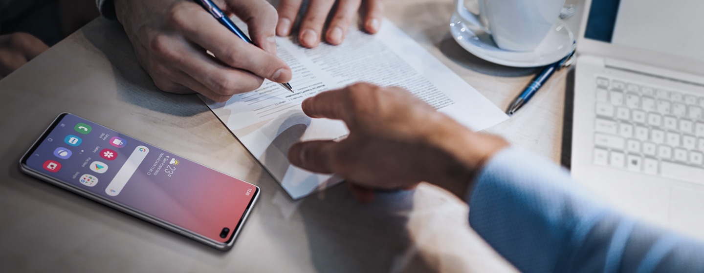 Imagen de un trabajador financiero mostrando algo de un documento a su cliente que está a punto de firmar el documento. El Galaxy S10 Plus está acostado a la izquierda en la mesa.