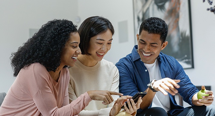 Una usuaria de Galaxy está sentada al lado de sus amigos y les muestra su teléfono inteligente con alegría mientras su amiga apunta hacia la pantalla del teléfono inteligente.