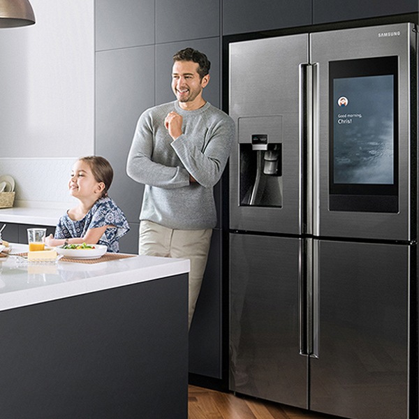 The man is standing in front of the refrigerator and the child is sitting at the table eating and laughing.
