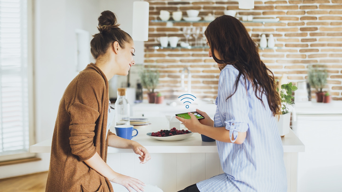 Two women sitting next to the kitchen table are watching TV show on the mobile phone. The Wi-Fi icon floating above her mobile phone means it's connected to the Samsung Smart TV.