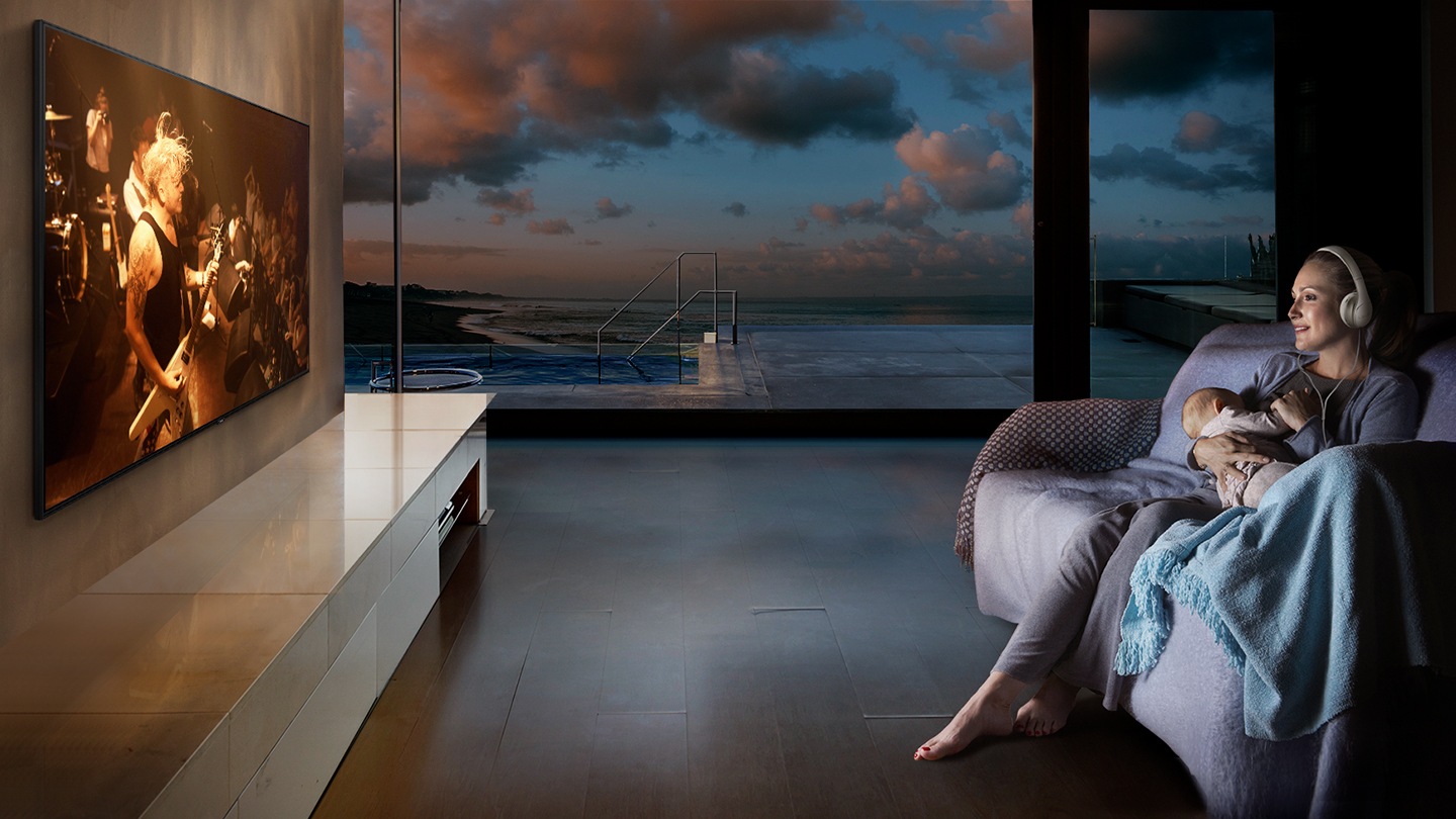A woman is watching TV in the living room with a headset while holding a baby at night.