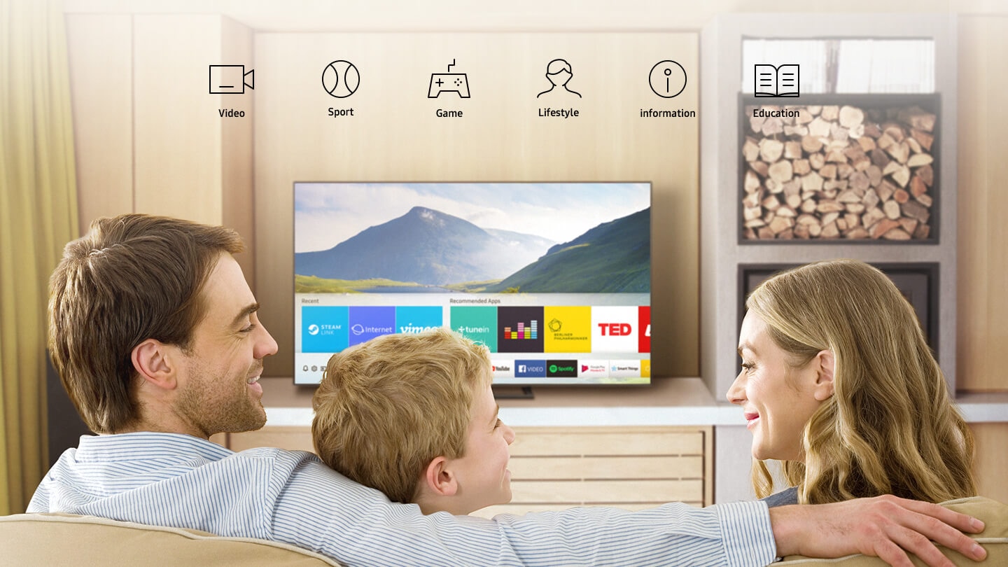 3 people in the living room are watching Smart TV. Smart Hub UI is displayed over a nature background image on TV screen; The UI contains various content, such as, Steam Live, Internet, Vimeo on 'Recent', Tunein Radio, Deezer, Berliner Philharmoniker, TED on 'Recommended Apps' and several applications like YouTube, Facebook Video, Google Play Movies & TV, SmartThings.