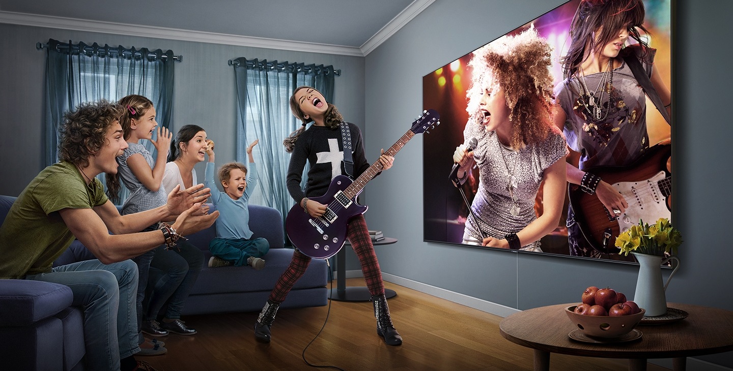A teenage girl jams along on her guitar to a rock concert playing on the Samsung Super Big TV, surrounded by cheering family members sitting around the living room.