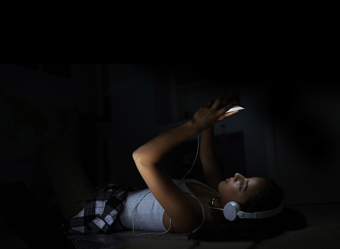 On voit une femme avec des écouteurs allongée sur un canapé, dans l’obscurité. Elle regarde l’écran brillant de son téléphone.
