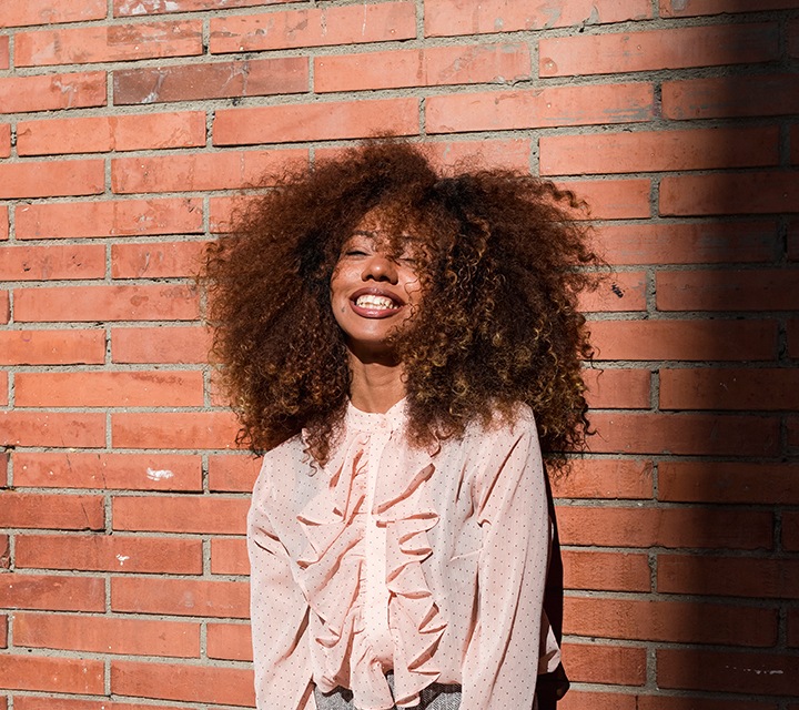 On voit un portrait d’une jeune femme souriante devant un mur en briques.