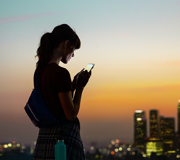 Een vrouw die bij zonsondergang op haar smartphone kijkt terwijl zij buiten staat met wolkenkrabbers op de achtergrond