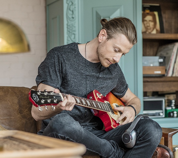 Man die thuis op zijn gitaar speelt met een AKG koptelefoon over zijn knie