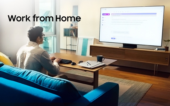 We zien een man op de bank in zijn woonkamer. Hij typt op een toetsenbord dat is aangesloten op zijn Samsung Smart TV. Hierop wordt het beeldscherm van zijn computer op kantoor weergegeven met behulp van Remote Access. Hij typt een e-mail en deze wordt weergegeven op het scherm