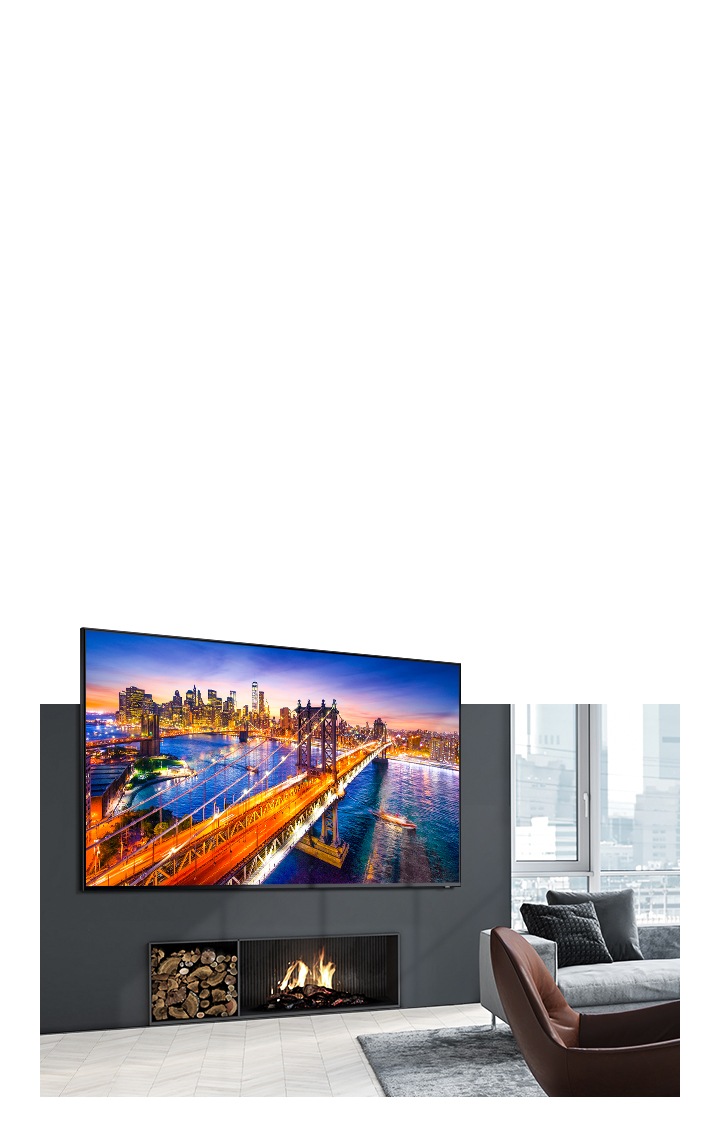 Een TV hangt aan de muur boven een open haard in een lichte en gezellige woonkamer en toont een nachtscène van een stad met een brug in heldere beeldkwaliteit dankzij de beeldinstellingen van Samsung Dynamic Mode.
