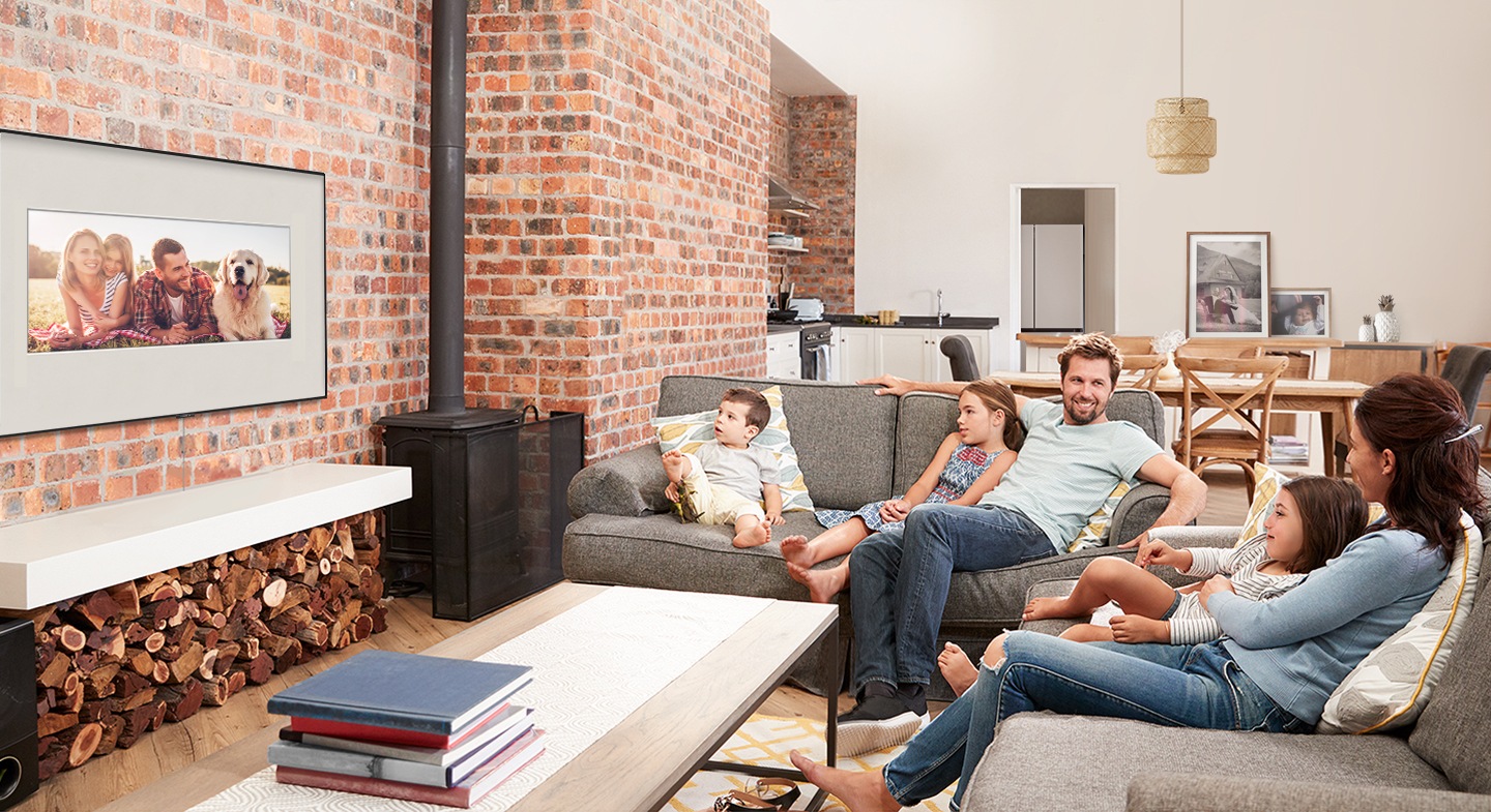 A family on sofa in the living room is watching family photos displayed on a wall with Samsung Ambient Mode to enjoy family memories.