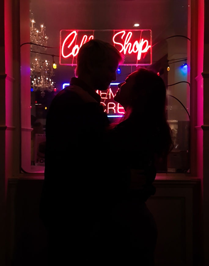 Silhouette photography of a couple standing in front of a neon coffee shop light, the scene is dark but you can make out their outline