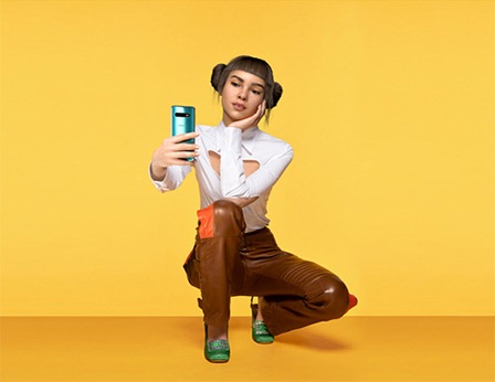 Primer plano de Lil Miquela agachada, sobre un fondo amarillo pálido, sosteniendo un teléfono inteligente Samsung Galaxy S10 azul espejado