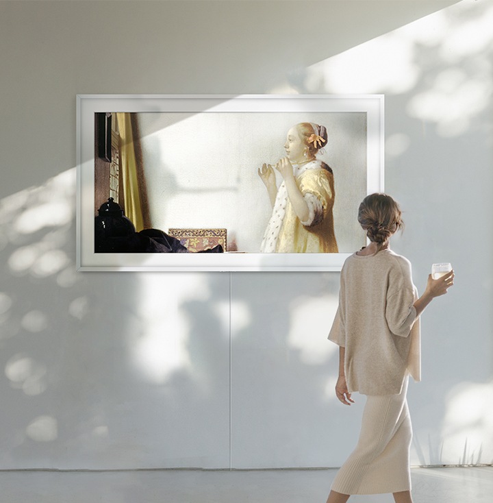The Frame on the wall is displaying one of Johannes Vermeer paintings, 'Woman with a Pearl Necklace', which makes living room a gallery, and a woman is watching the artwork.