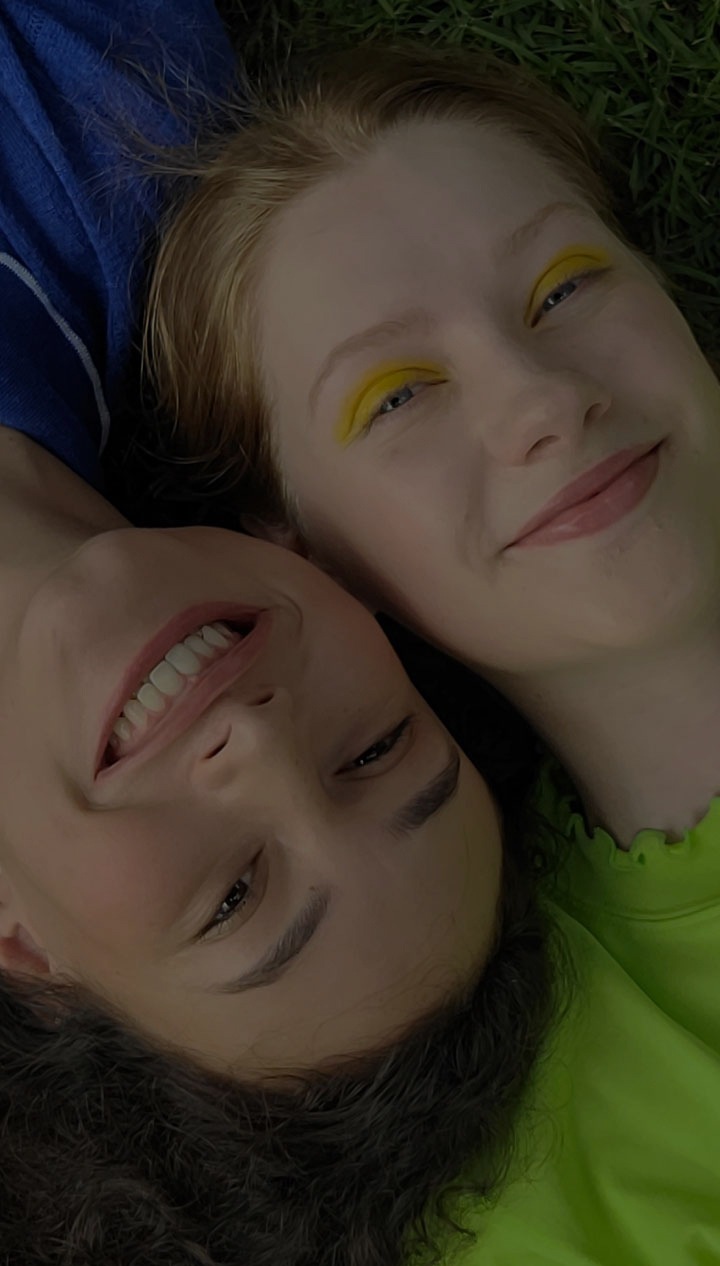 An aerial view of two women laying side-by-side on the grass. The woman on the left is wearing a blue collared shirt while the woman on the right is wearing a lime green top. Their heads are pressed together from opposite ends. They're both smiling at the camera.