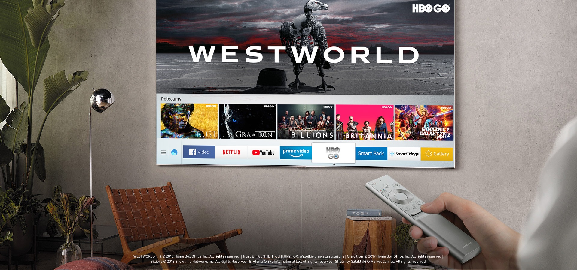 Różnorodność kanałów wyświetlonych na nowym telewizorze Samsung QLED TV, który jest zawieszonym na ścianie oraz ręka mężczyzny trzymająca pilota.