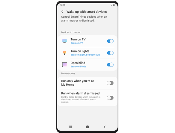 Um ecrã do smartphone Galaxy a mostrar a página de configurações 'Despertar com dispositivos inteligentes'. Em 'Dispositivos a controlar', está escrito "Ligue a TV", "Ligue as luzes" e "Abra as cortinas". Todos os três comandos estão ativados e por baixo de cada comando lê-se, respetivamente, "TV do quarto", "Luz do quarto, Lâmpada do quarto" e "Cortinas do quarto". Abaixo de "Dispositivos para controlar", "Mais opções" fornecem as seguintes opções de configuração que estão atualmente desativadas: "Correr apenas quando estiver em Minha casa" e "Correr quando o alarme for desligado".