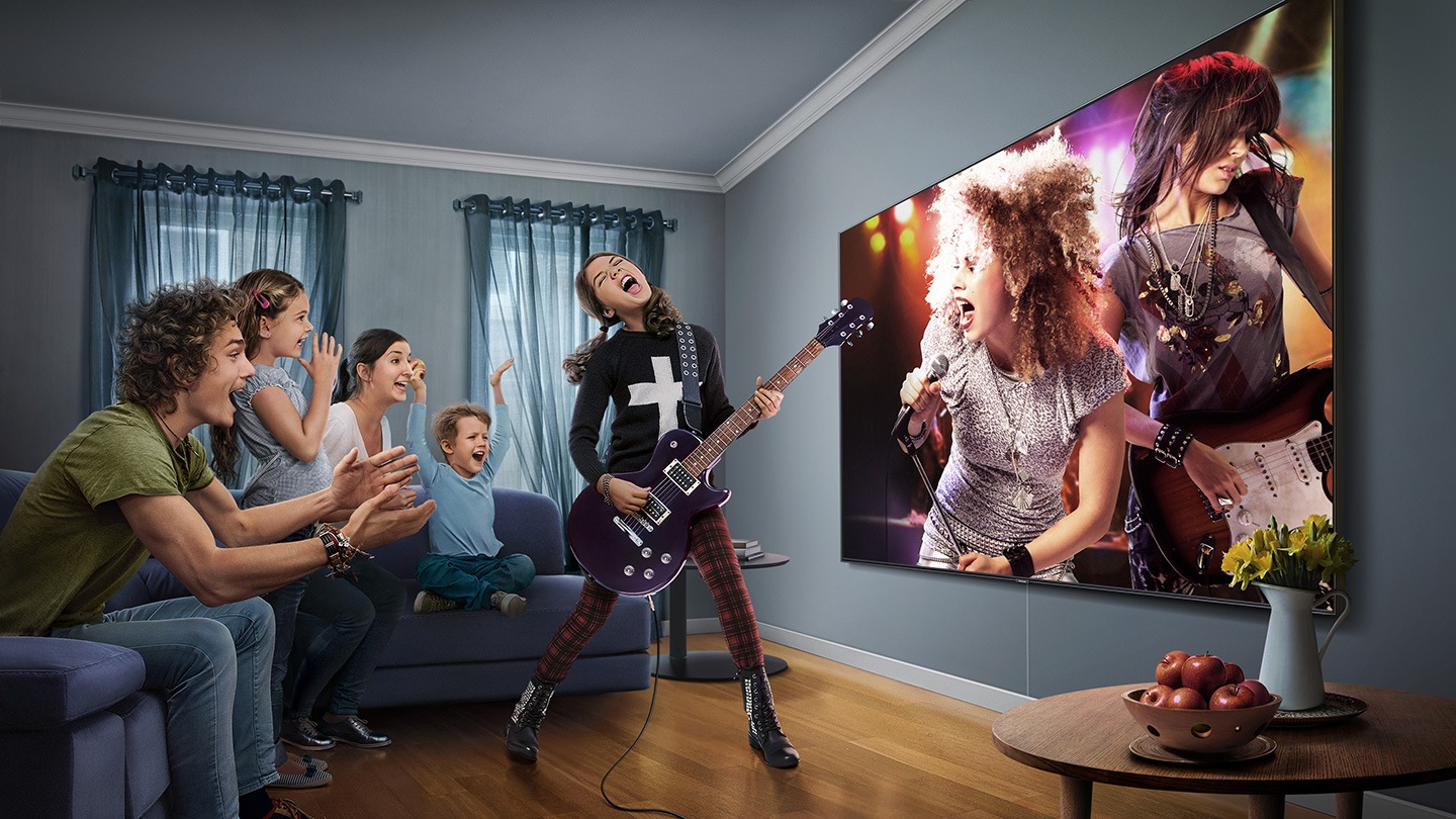 Super Big TV Cinco pessoas reunidas numa sala a assistir a um concerto numa televisão com um grande ecrã. Quatro estão sentadas no sofá a aplaudir enquanto uma rapariga toca guitarra elétrica no centro da sala.